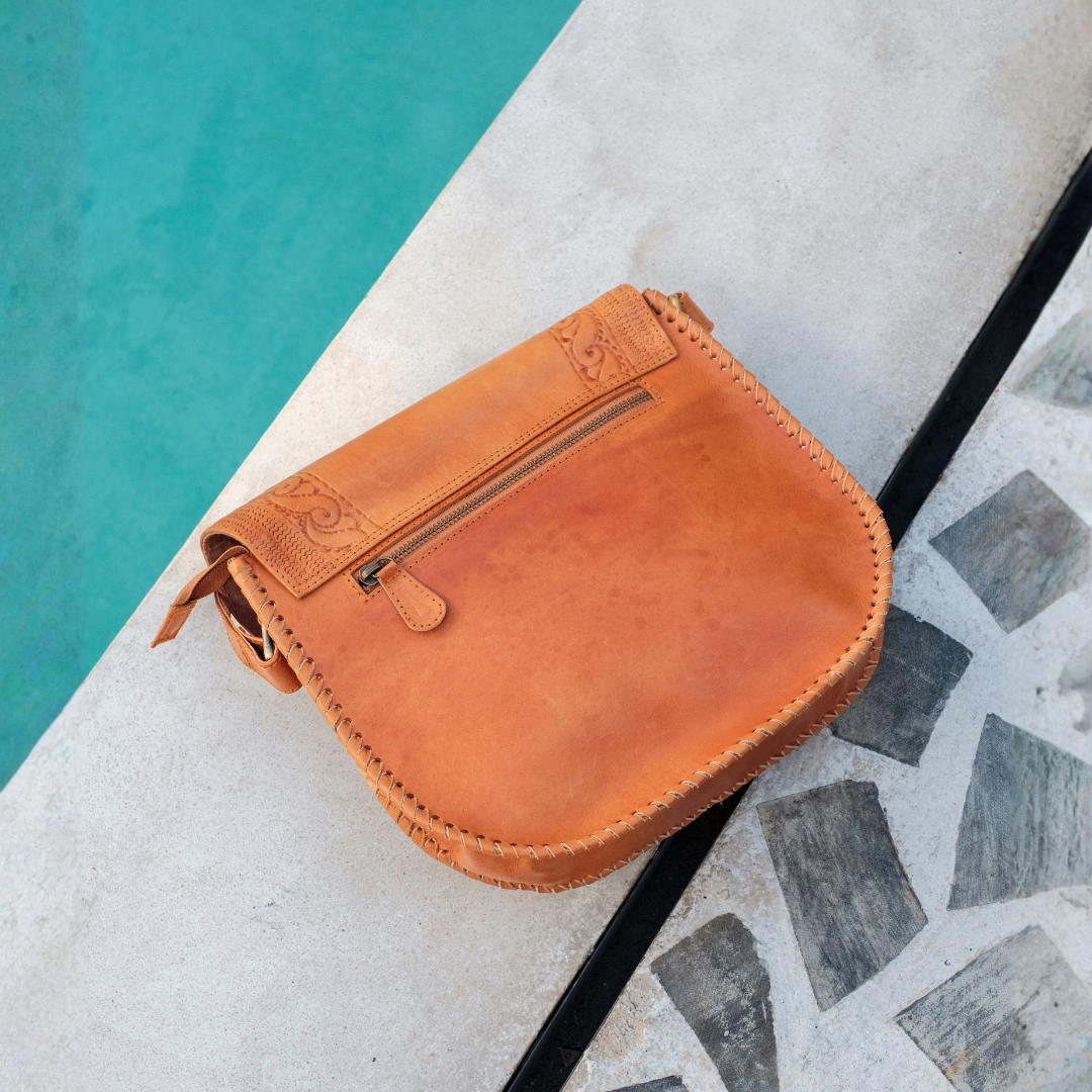 Saddler Crossbody Bag from House of Hide in tan leather with front zipper pocket being displayed on concrete surface next to turquoise pool