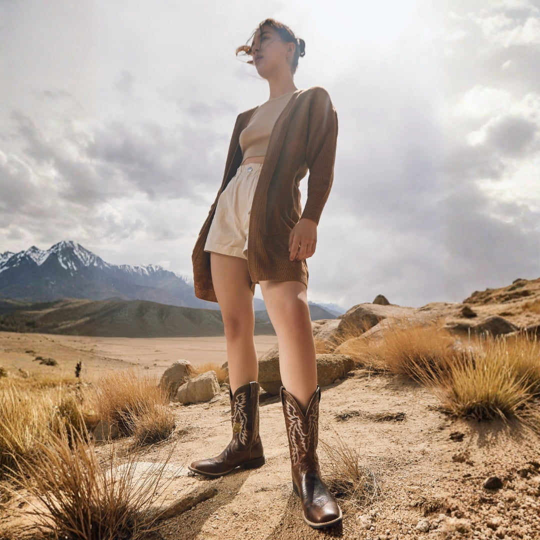 Model wearing Salvage brown leather cowboy boots with cream shorts and a brown cardigan in a mountain landscape.