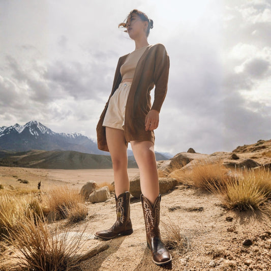 Model wearing Salvage brown leather cowboy boots with cream shorts and a brown cardigan in a mountain landscape.