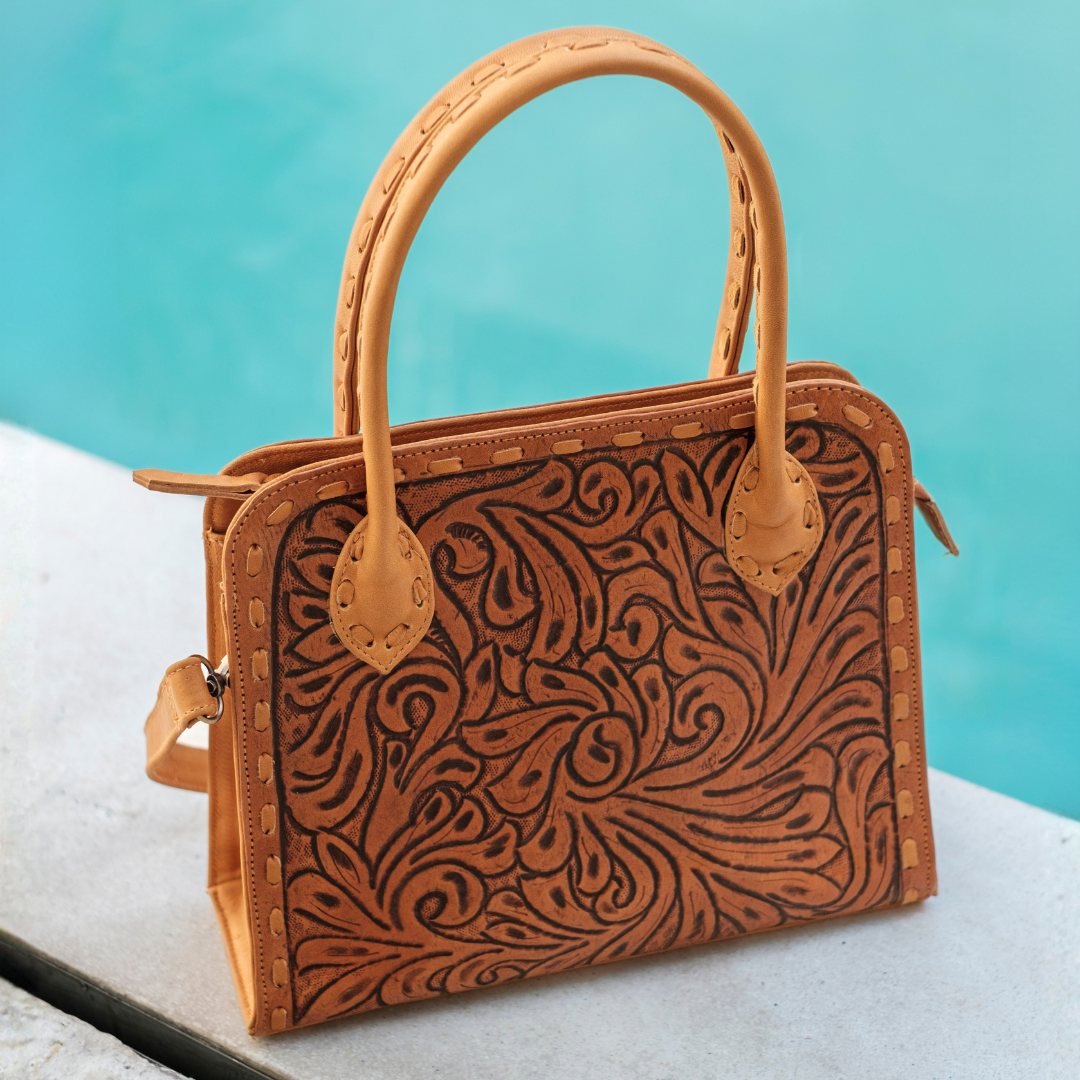 A tan tooled leather handbag with intricate swirling floral patterns, featuring dual top handles and a zip-around closure, set against a turquoise pool background.