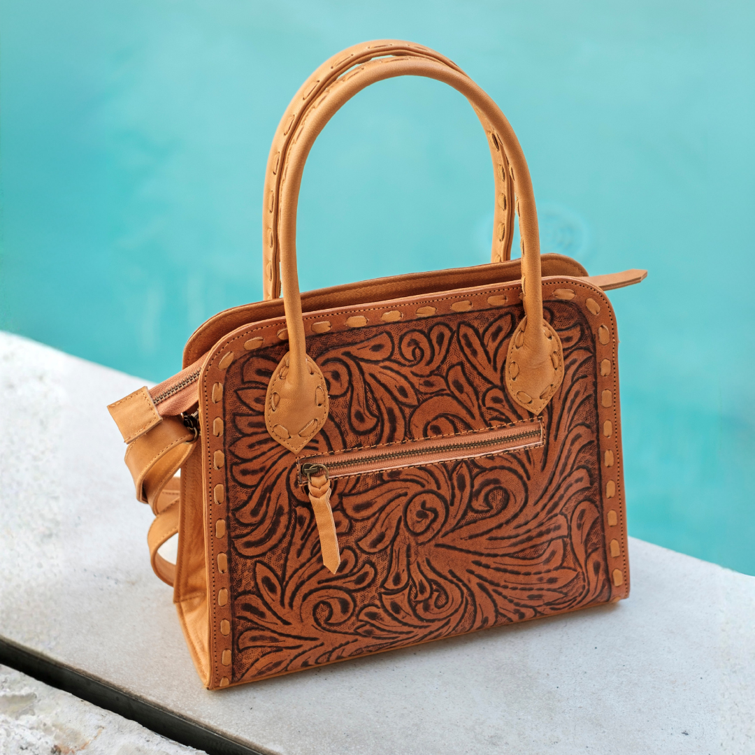A structured tan leather handbag with an embossed floral scroll pattern, dual top handles, and a front zip pocket, photographed on a light stone surface with a turquoise water background.