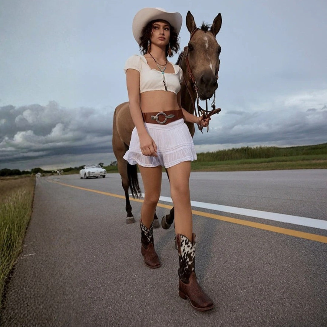 Model wearing Savanna cowhide cowboy boots with white shorts and a cream hat, leading a horse.