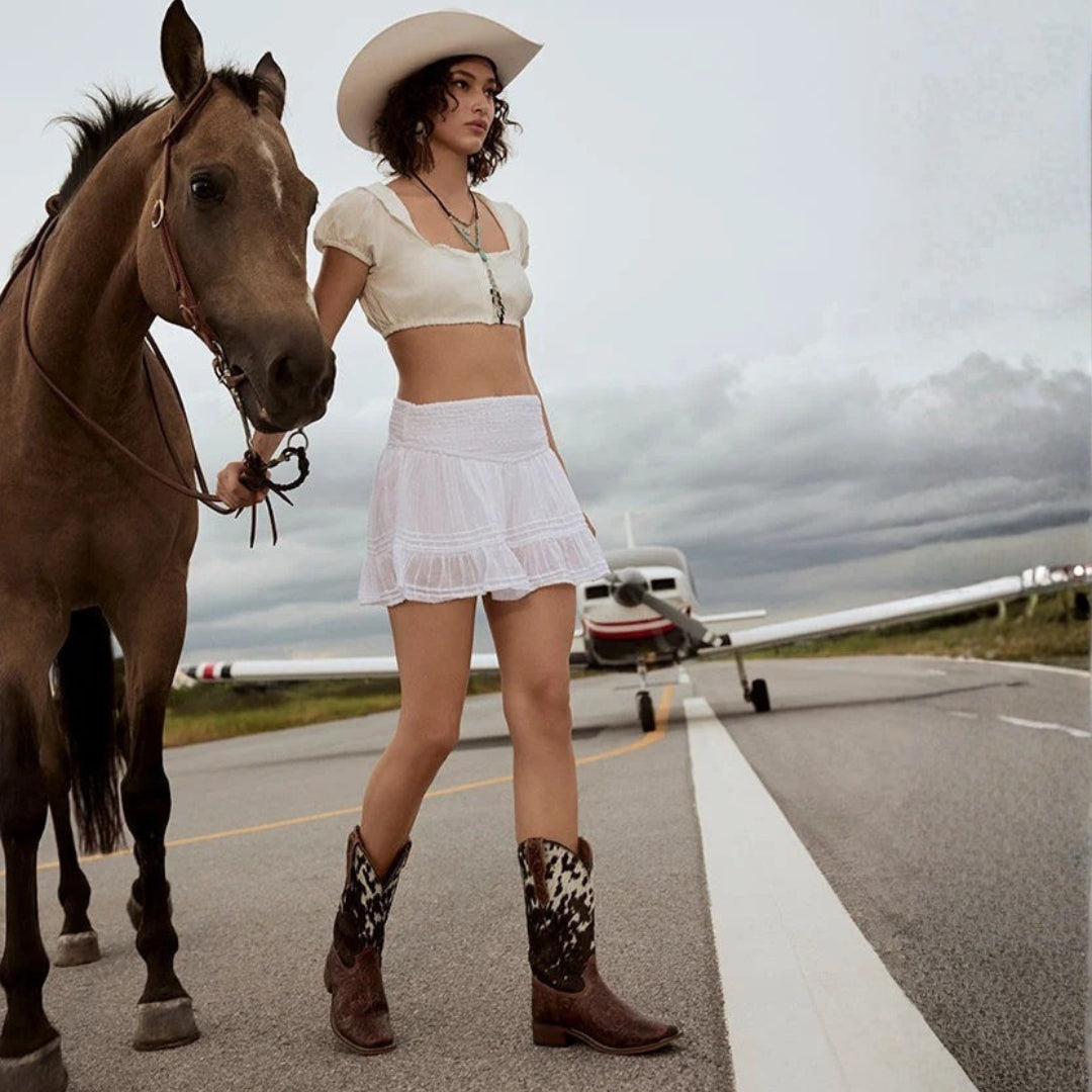 Savanna leather cowboy boots styled with a white outfit beside a horse, with a light plane behind.