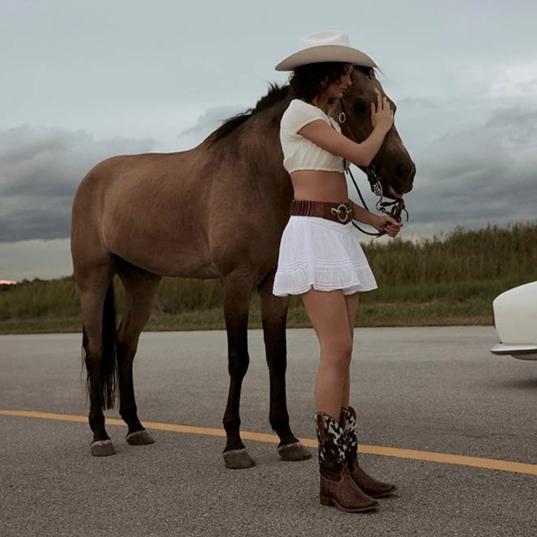 Side view of Savanna brown leather and cowhide boots worn by a model standing beside a horse.