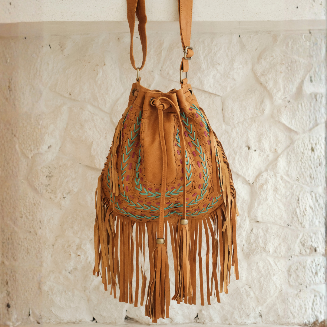Sedona Drawstring Bag from House of Hide in tan leather with fringe detail, turquoise and brown embroidered detailing, and drawstring closure being displayed hanging against textured white wall