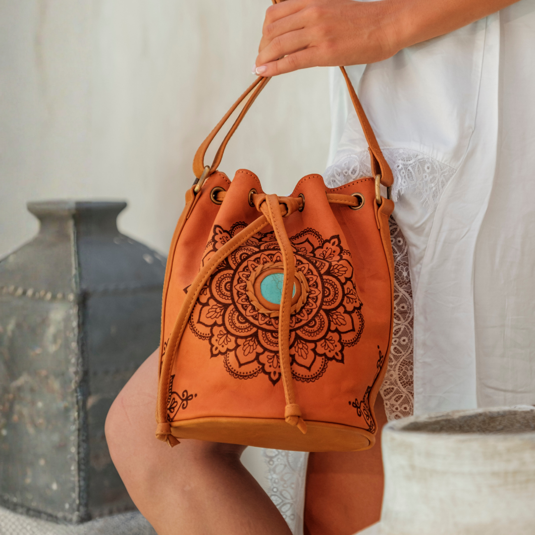 Soleil Mandala Bucket Bag from House of Hide in tan leather with large brown mandala print and turquoise stone center being held by model with dark metal container in background