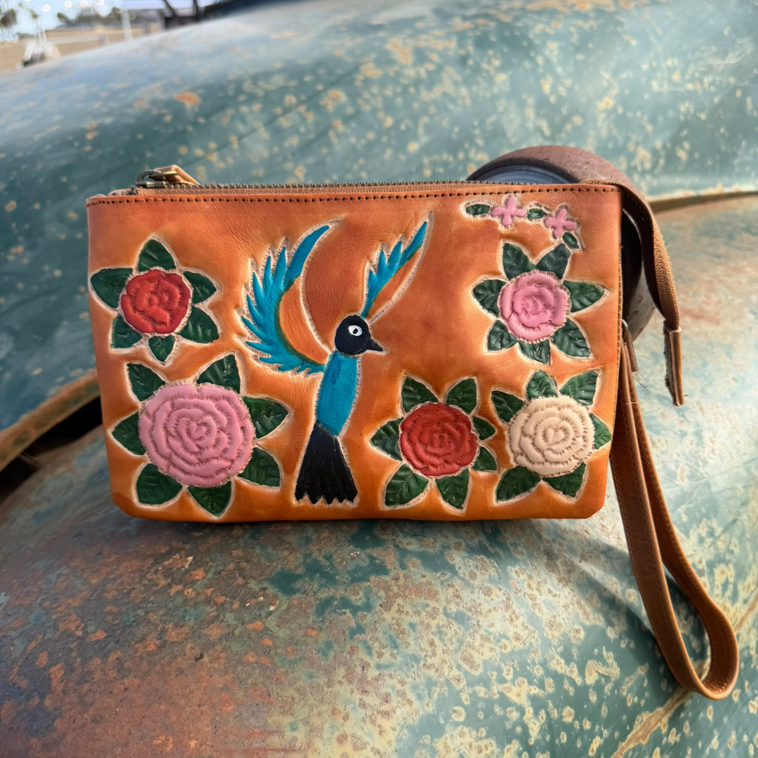 A tan leather wristlet clutch with hand-stitched floral and bird embroidery, resting on a weathered green metal surface.