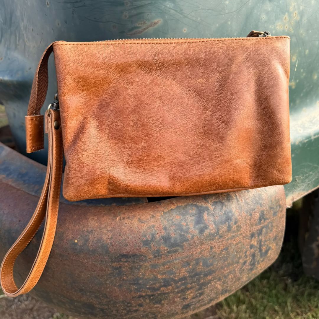 A tan leather wristlet clutch with a top zipper and wrist strap, resting on a rustic metal surface outdoors.