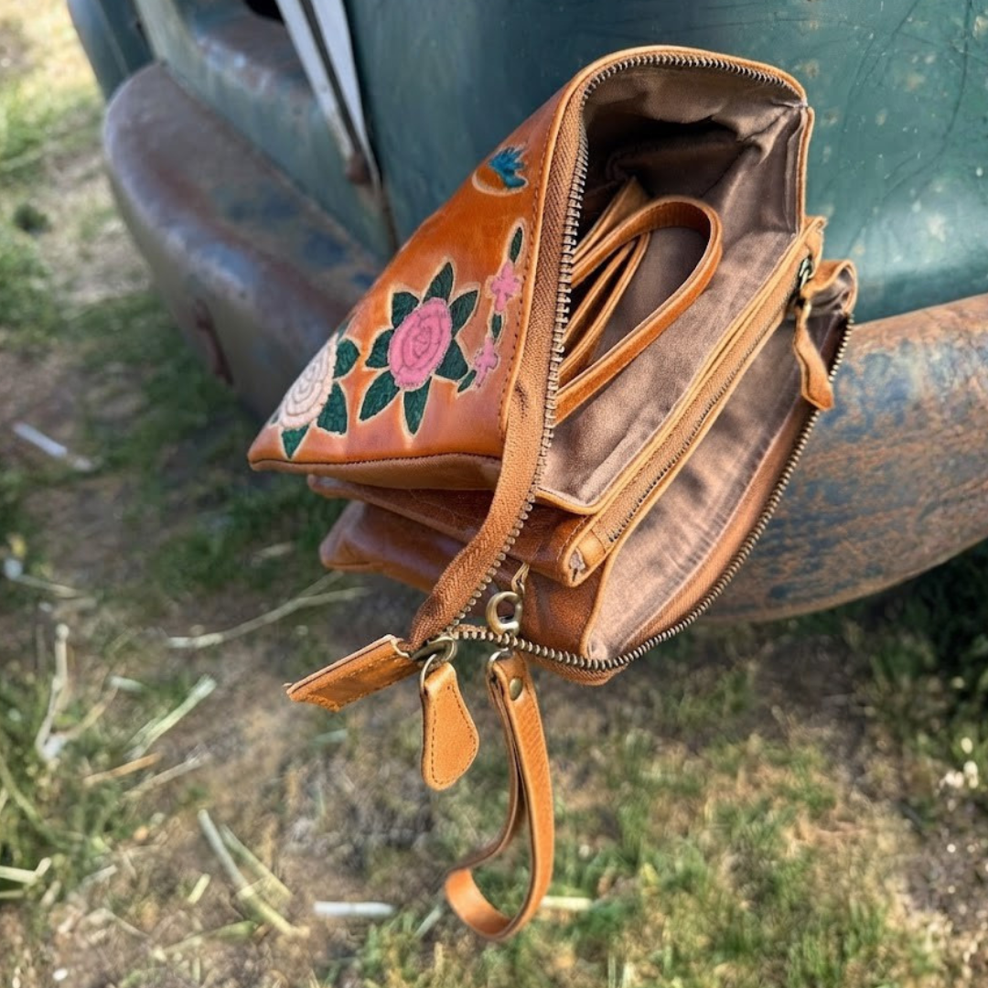 A brown leather crossbody bag with hand-painted pink and green floral embroidery, shown open and resting against a weathered green vehicle outdoors.