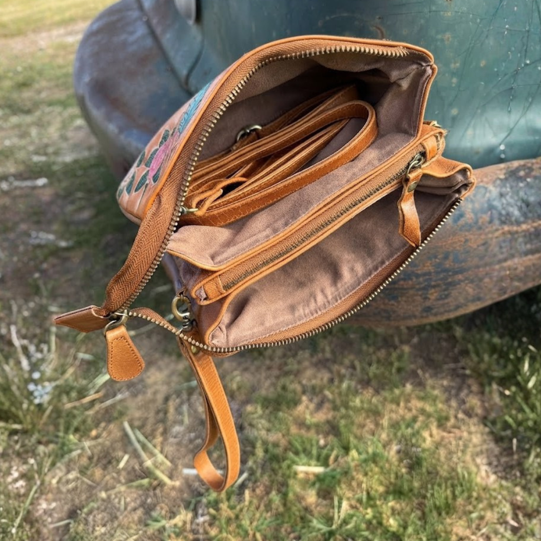 A tan leather crossbody bag with floral embroidery on the exterior, shown open to display its interior compartments, lining, and strap. The bag is photographed outdoors on grass next to a large metal sculpture.
