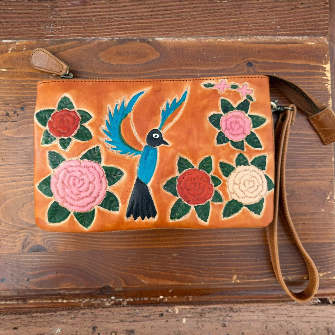 An orange leather wristlet clutch with hand-painted floral and bird motifs, displayed on a rustic wooden surface.