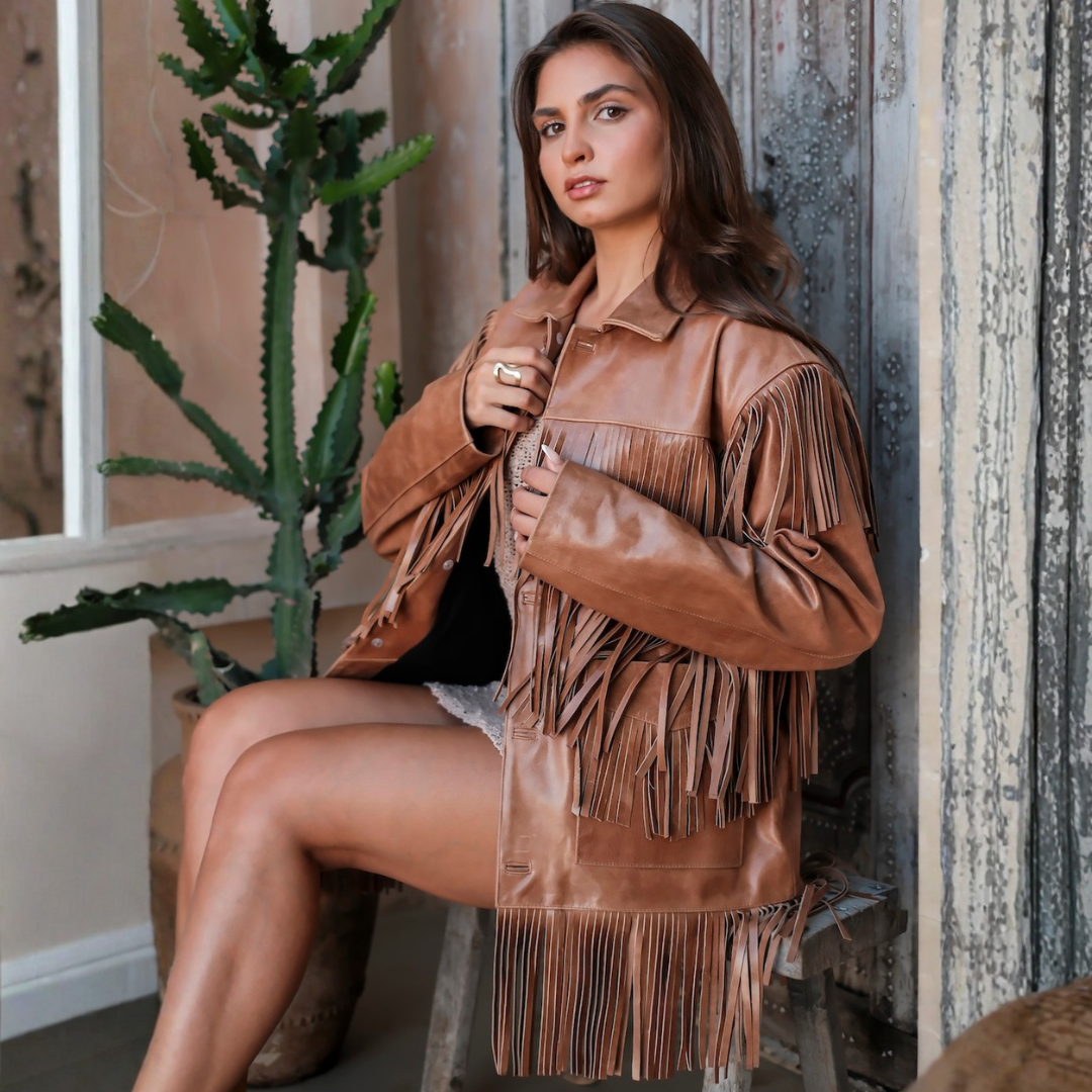 A woman wearing a tan faux-leather fringe jacket sits on a stool in a rustic indoor setting with a cactus plant in the background.
