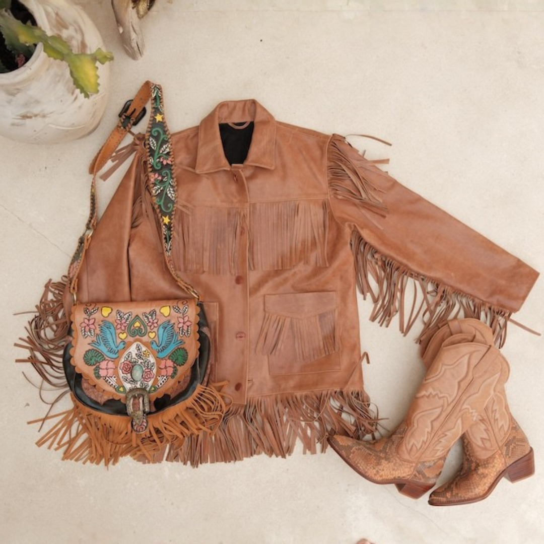 A styled flat-lay of western wear featuring a tan leather fringe jacket, a matching embroidered shoulder bag, and a pair of tan leather cowboy boots on a light beige surface.