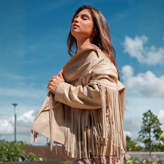 A woman wearing a beige fringe jacket with eyelet detailing, photographed outdoors against a blue sky with clouds.
