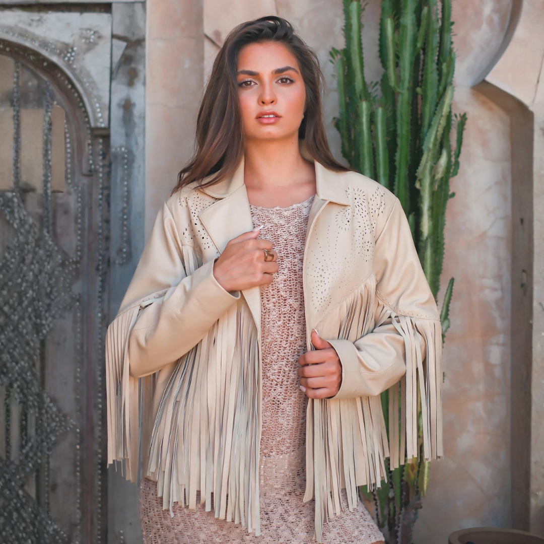 A woman wearing a beige fringe jacket over a matching lace dress, standing in front of a stone wall and cactus.