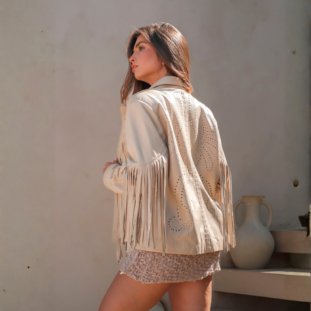 A woman wearing a beige fringed sleeveless jacket with laser-cut circular patterns, photographed from behind against a neutral beige wall.
