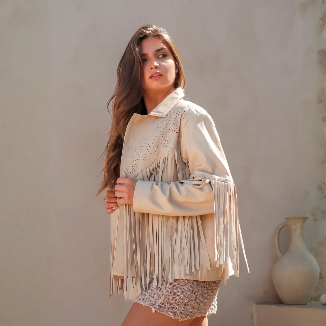 A woman wearing a cream-colored fringe jacket with eyelet embroidery on the chest, posed against a neutral beige wall with a ceramic vase in the background.