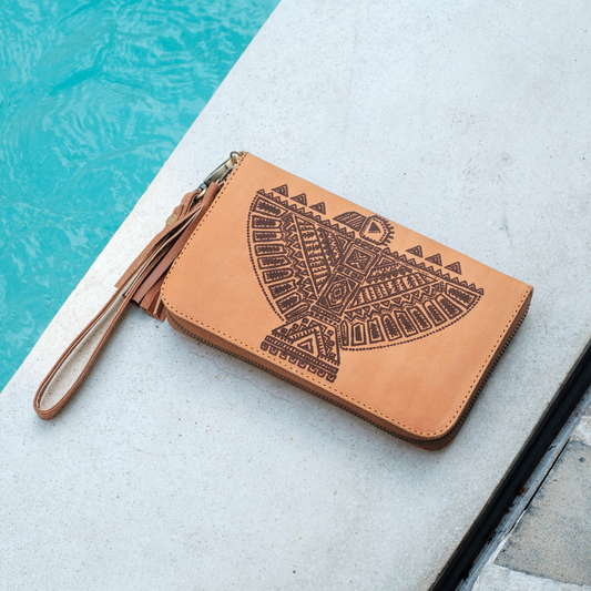 Thunderbird Wristlet Pouch from House of Hide in tan leather with dark brown geometric eagle design and wrist strap being displayed on light concrete near swimming pool