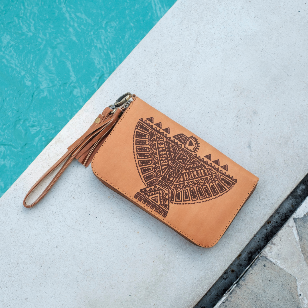 Thunderbird Wristlet Pouch from House of Hide in light tan leather with printed geometric eagle design and tassel being displayed on concrete surface next to turquoise pool
