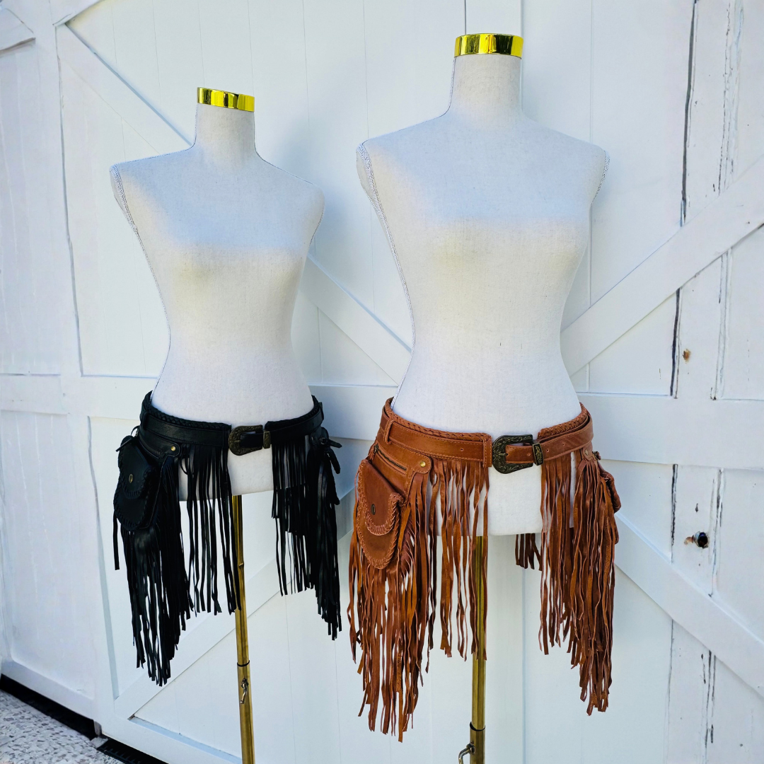 Two faux leather fringe waist belts displayed on dress forms, one in black and one in brown, set against a white wooden barn-style background.
