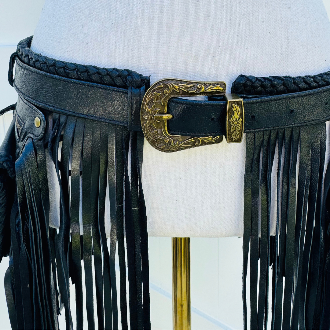 A black leather fringe belt with braided trim and an ornate antique gold buckle, displayed on a white dress form mannequin.