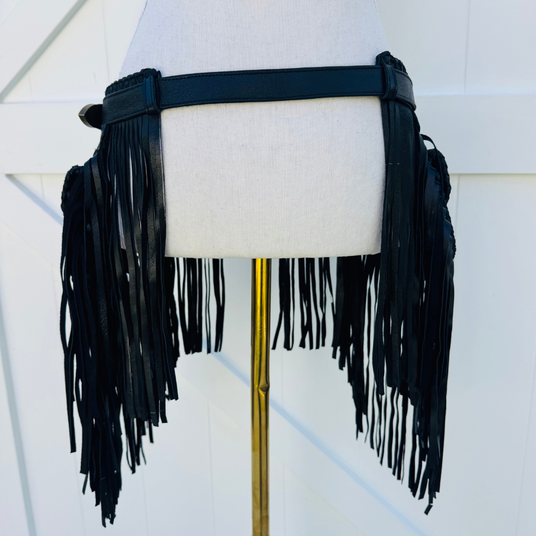A black faux-leather fringe belt with a wide buckle strap, displayed on a cream fabric mannequin torso stand with a gold pole against a white paneled background.