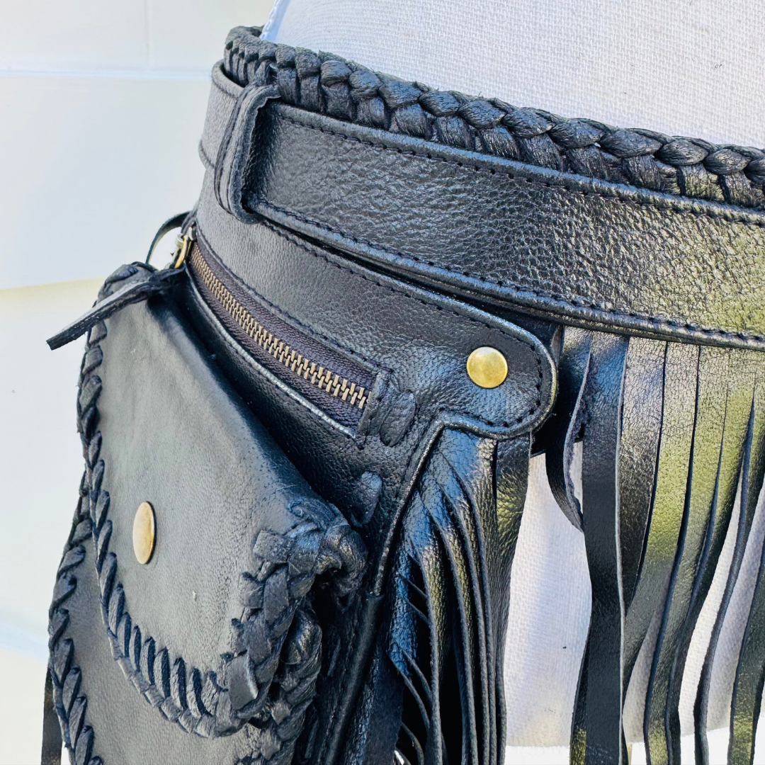 Black leather fringe belt bag with braided details, a zippered pocket, and gold-tone hardware, shown on a white mannequin torso.