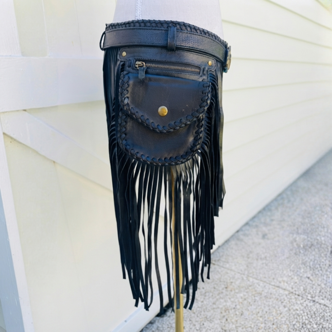 Black leather fringe pouch bag with braided trim and a top zipper, shown hanging against a white exterior wall with plants in the background.
