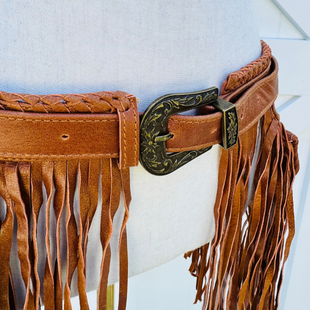 A close-up of a brown leather fringe belt with braided detailing and an ornate engraved metal buckle, displayed on a white dress form.