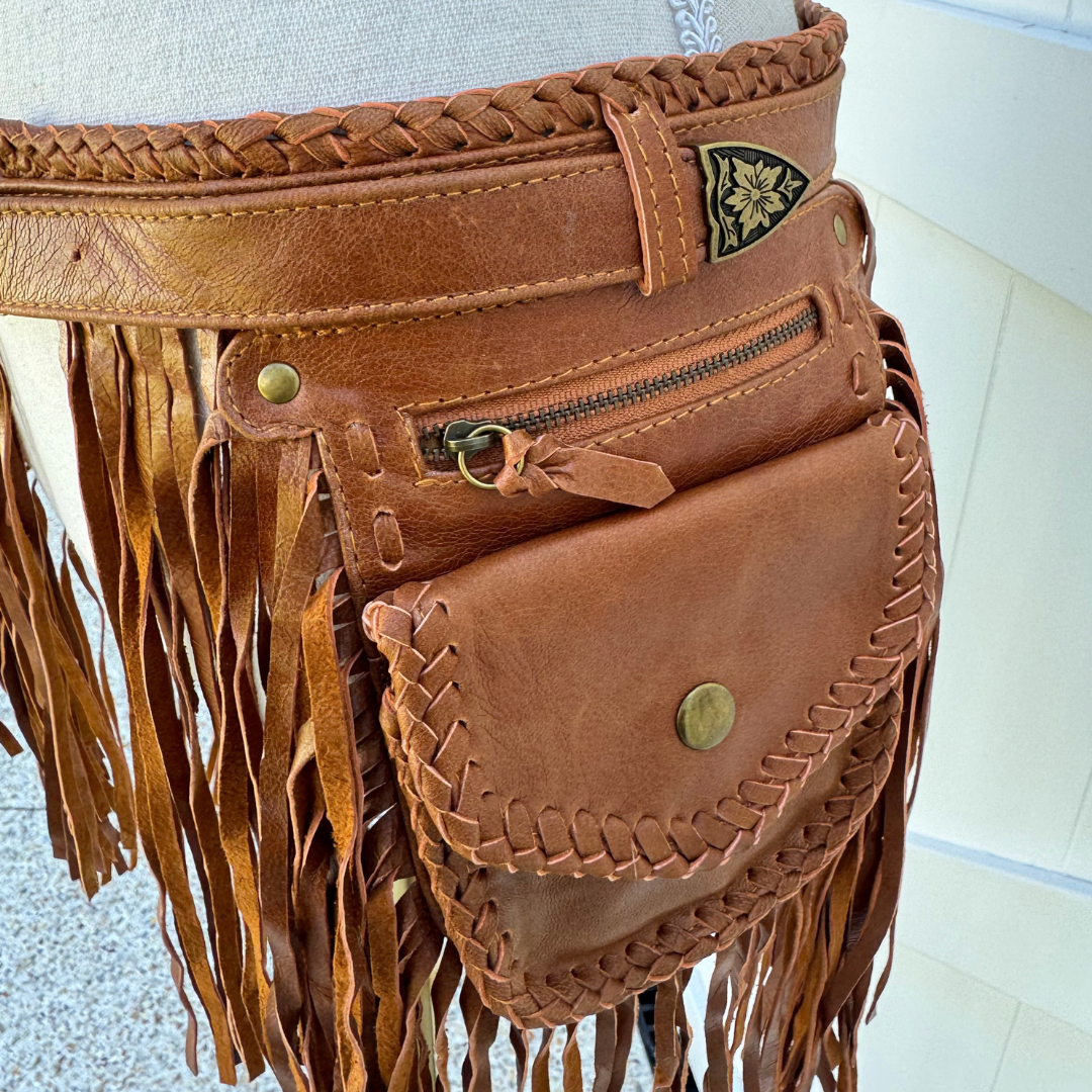 A close-up of a brown leather belt bag with a wide braided waist belt, long fringe, a zippered pocket, and a flap pocket with brass-tone hardware, displayed on a mannequin.