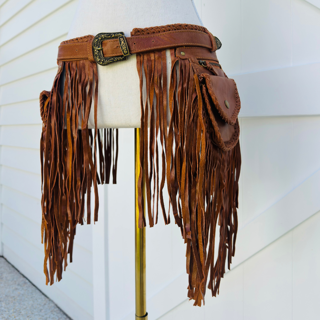 A brown leather fringe belt with a large decorative buckle and a side pouch, displayed on a cream dress form mannequin against a white siding background.