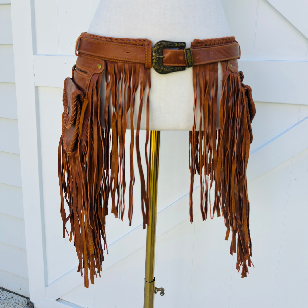 A brown leather fringe belt with multiple utility pockets and a dark metal buckle, displayed on a cream dress form mannequin stand against a white siding background.