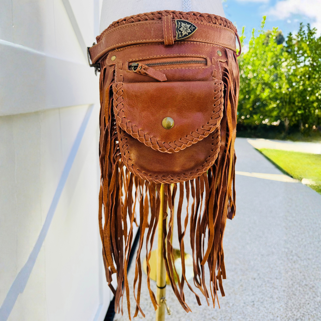 A brown leather fringe pouch bag with braided details and a front pocket, displayed on a stand outdoors next to a white wall.