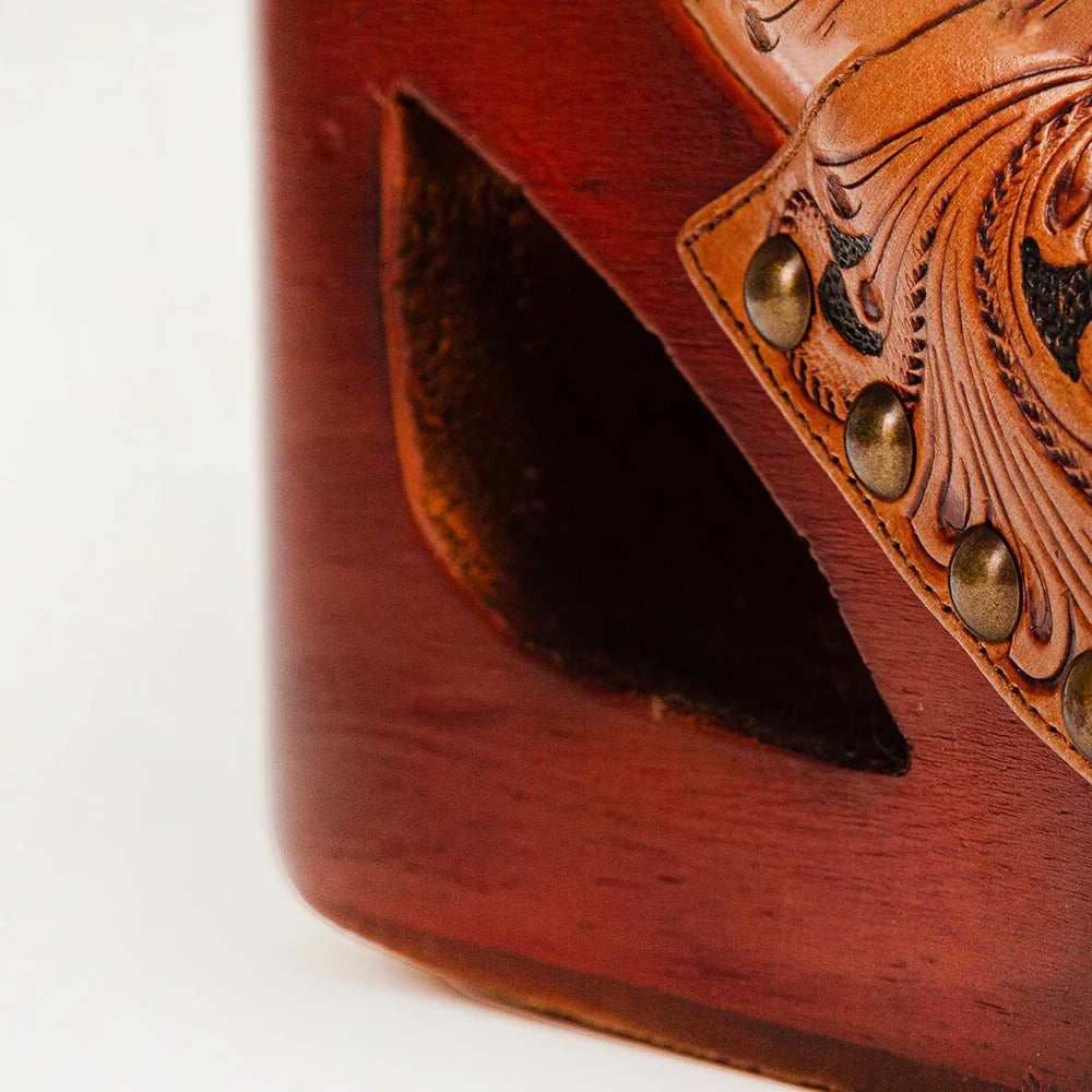 Close-up of a reddish-brown wooden object with a carved leather detail featuring brass-tone rivets and floral tooling, photographed against a plain light background.