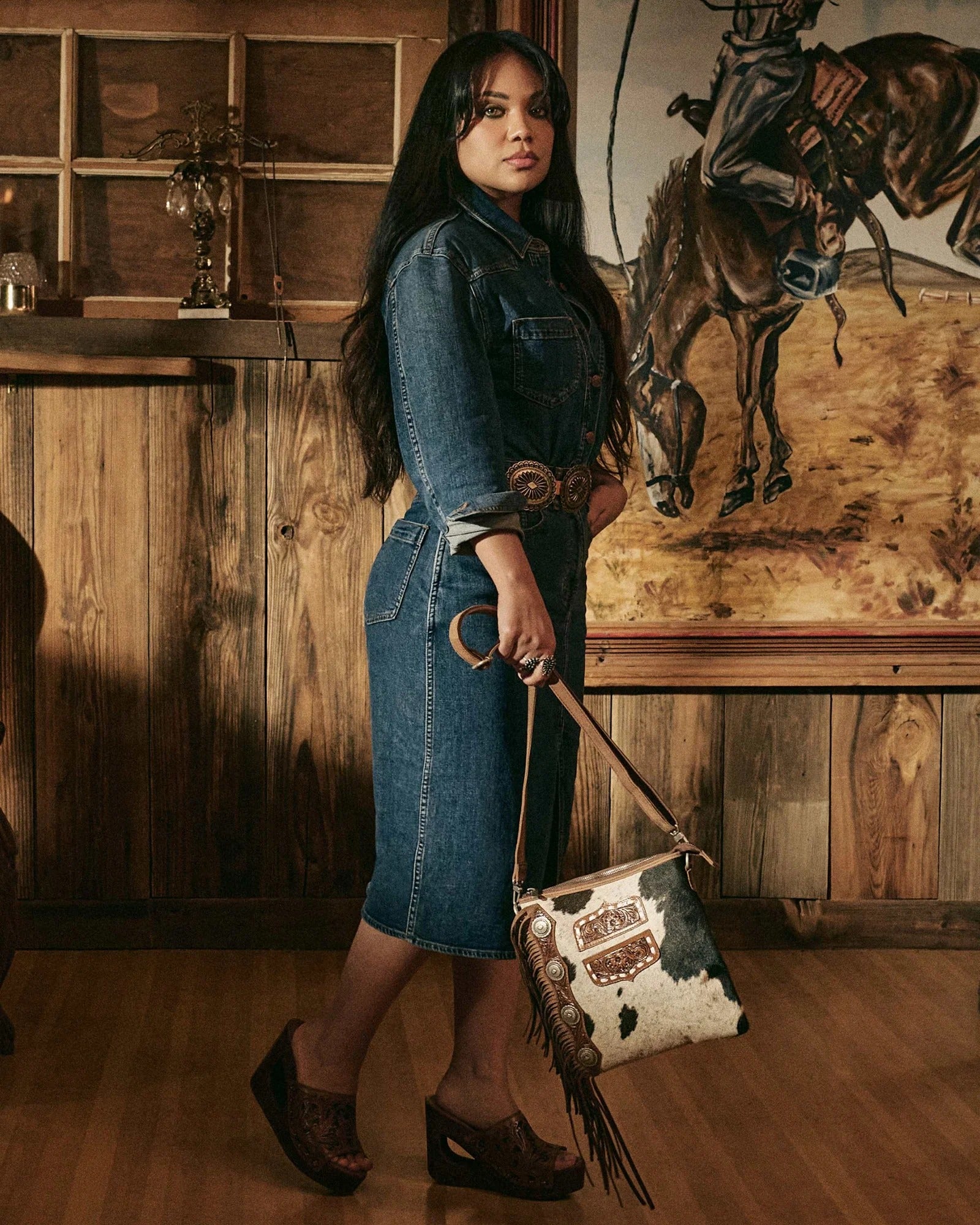 Model showcasing House of Hide women's western fashion wearing a denim dress and carrying a genuine leather cowhide handbag in a rustic setting