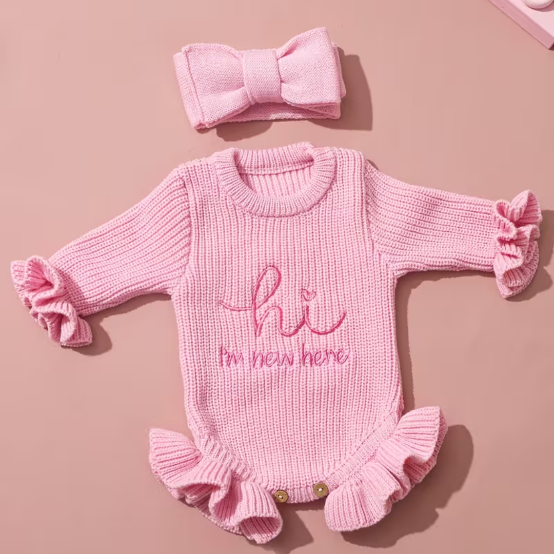 Welcome Baby Romper Set from House of Hide in soft pink chunky ribbed knit with hi I'm new here embroidery, ruffled cuffs and hem, snap button closure, matching bold bow headband being displayed on round woven mat for newborn photography