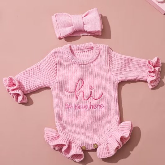 Welcome Baby Romper Set from House of Hide in soft pink chunky ribbed knit with hi I'm new here embroidery, ruffled cuffs and hem, snap button closure, matching bold bow headband being displayed on round woven mat for newborn photography