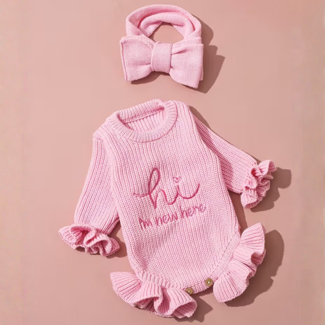 Welcome Baby Romper Set from House of Hide in soft pink ribbed knit long-sleeve romper with embroidered hi I'm new here text, ruffled edges, coordinating bow headband, photo prop set designed for precious first baby pictures on woven placemat