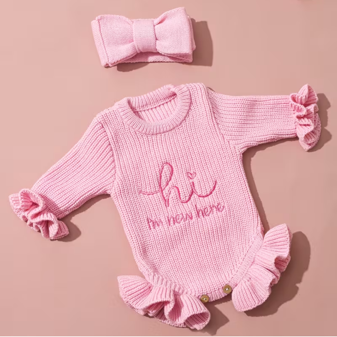 Welcome Baby Romper Set from House of Hide in delicate soft pink chunky ribbed knit romper with embroidered hi I'm new here announcement, picture-perfect ruffled edges, bold matching bow headband, snap button functionality on round woven display for photo shoots