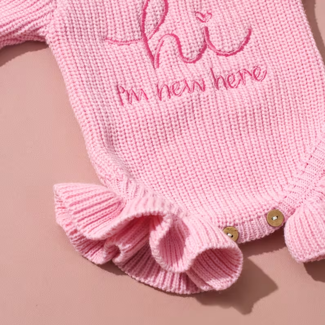 Welcome Baby Romper Set from House of Hide in soft pink ribbed knit with embroidered hi I'm new here text, gentle stretch construction, ruffled trim detail, bold bow headband that steals the show, snap button closure on circular woven mat backdrop