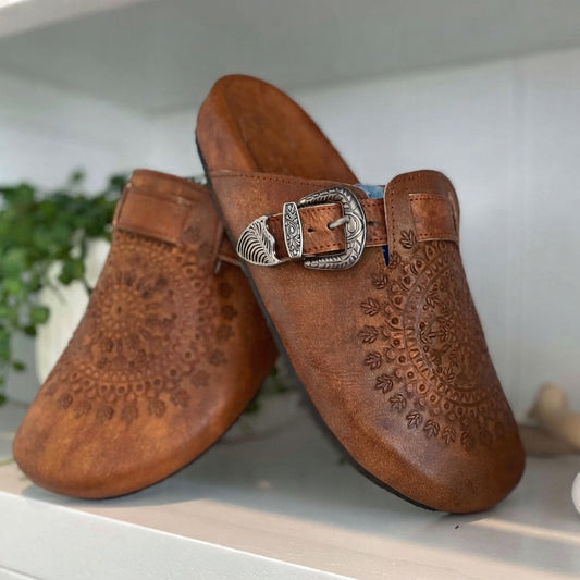 A pair of brown embossed leather clogs with a silver western-style buckle, displayed on a white shelf with a blurred green plant in the background.
