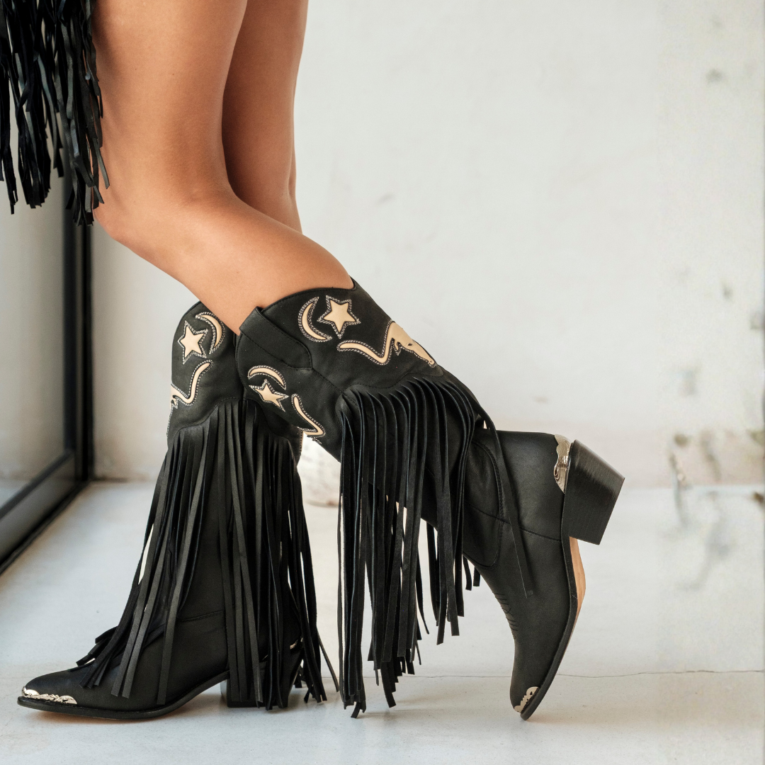 Black leather fringe cowboy boots with gold embroidered stars and western stitching, shown on a model's feet against a light gray marble floor.