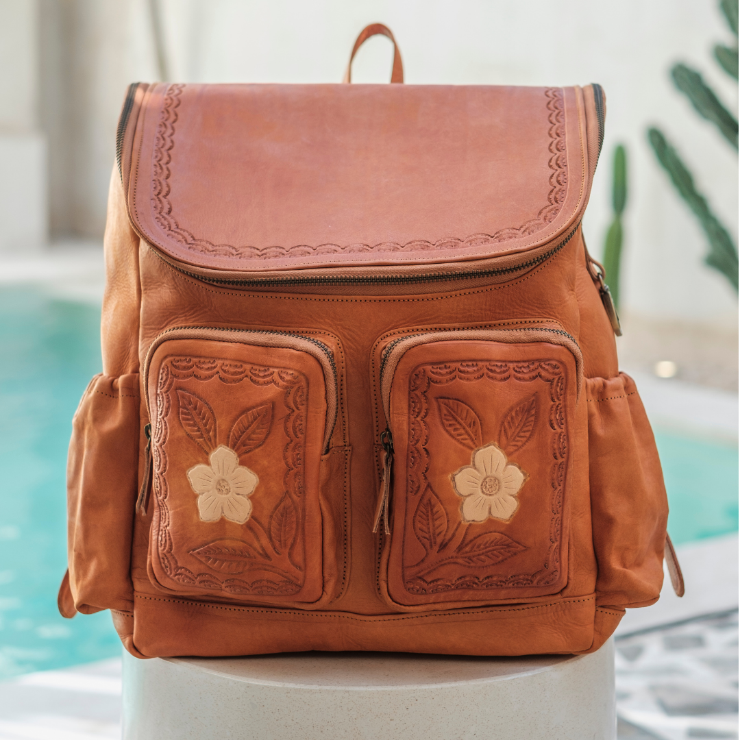 Wild Rose Leather Backpack from House of Hide in brown leather with embossed floral details on front pockets being displayed on white pedestal with poolside background
