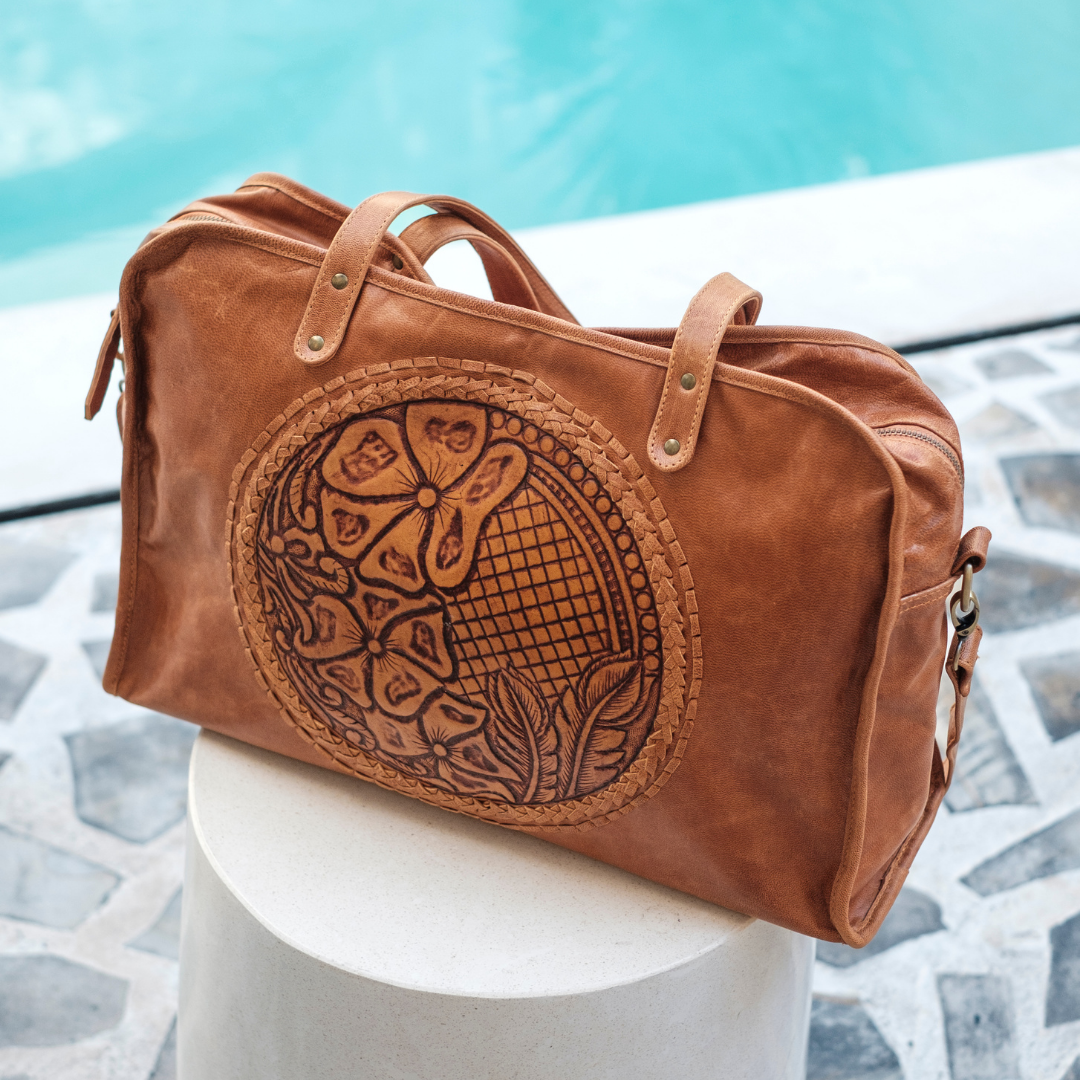 Wildflower Duffel Bag from House of Hide in brown leather with embossed floral design being displayed on white cylindrical pedestal in front of swimming pool