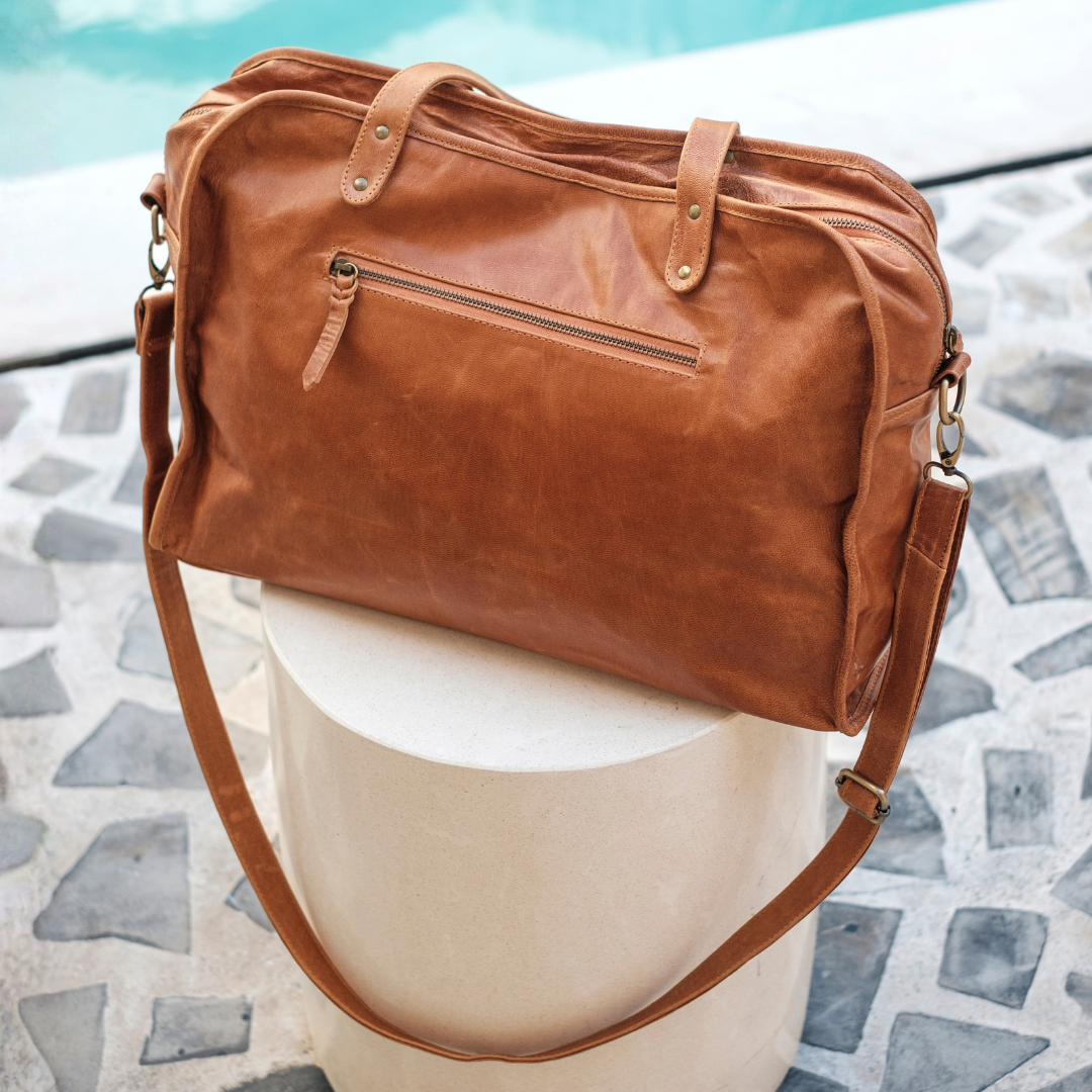 Wildflower Duffel Bag from House of Hide in brown leather with long shoulder strap being displayed on round white pedestal against poolside stone patio background