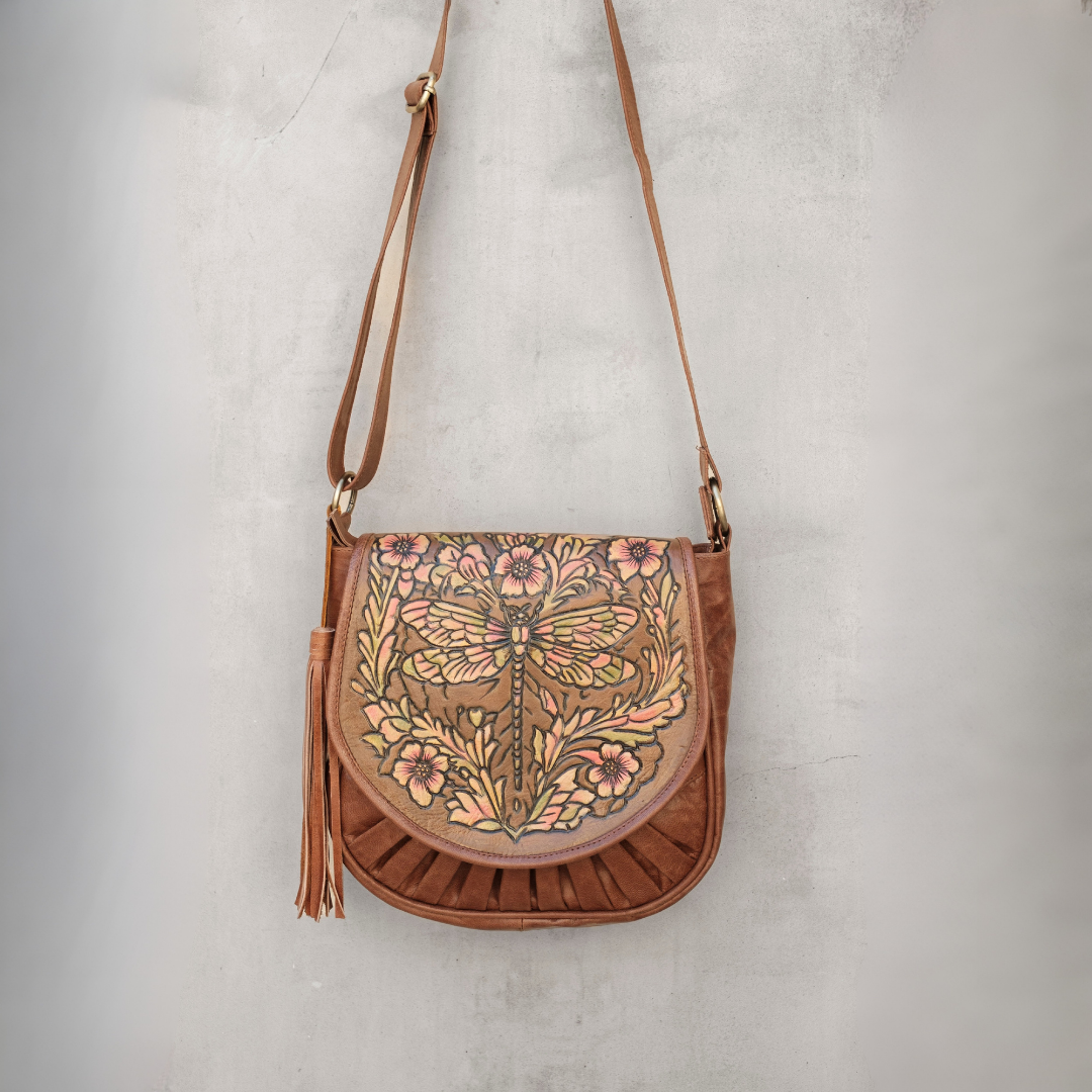 Willow Crossbody Bag from House of Hide in brown leather with tassel and detailed floral dragonfly design on front flap being displayed hanging against textured gray wall