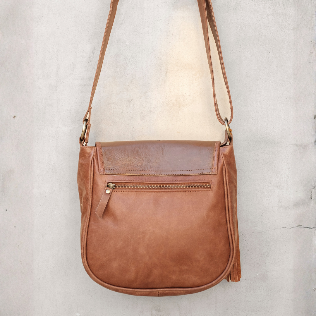 Willow Crossbody Bag from House of Hide in medium brown leather with front zippered pocket and matching shoulder strap being displayed against textured light gray concrete wall