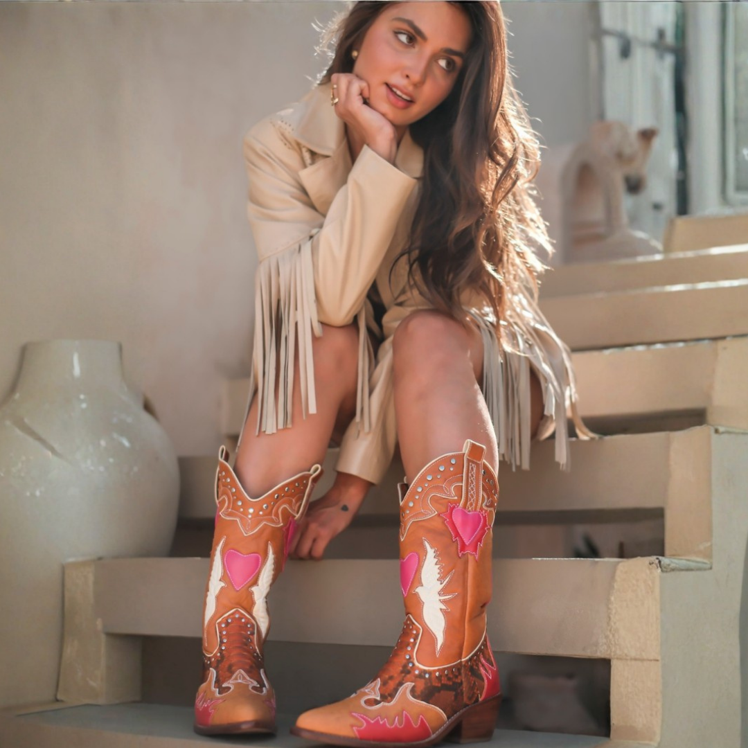 A woman sits on outdoor stone steps wearing a pair of hand-painted cowboy boots with orange, pink, and white designs, along with a beige fringe jacket.