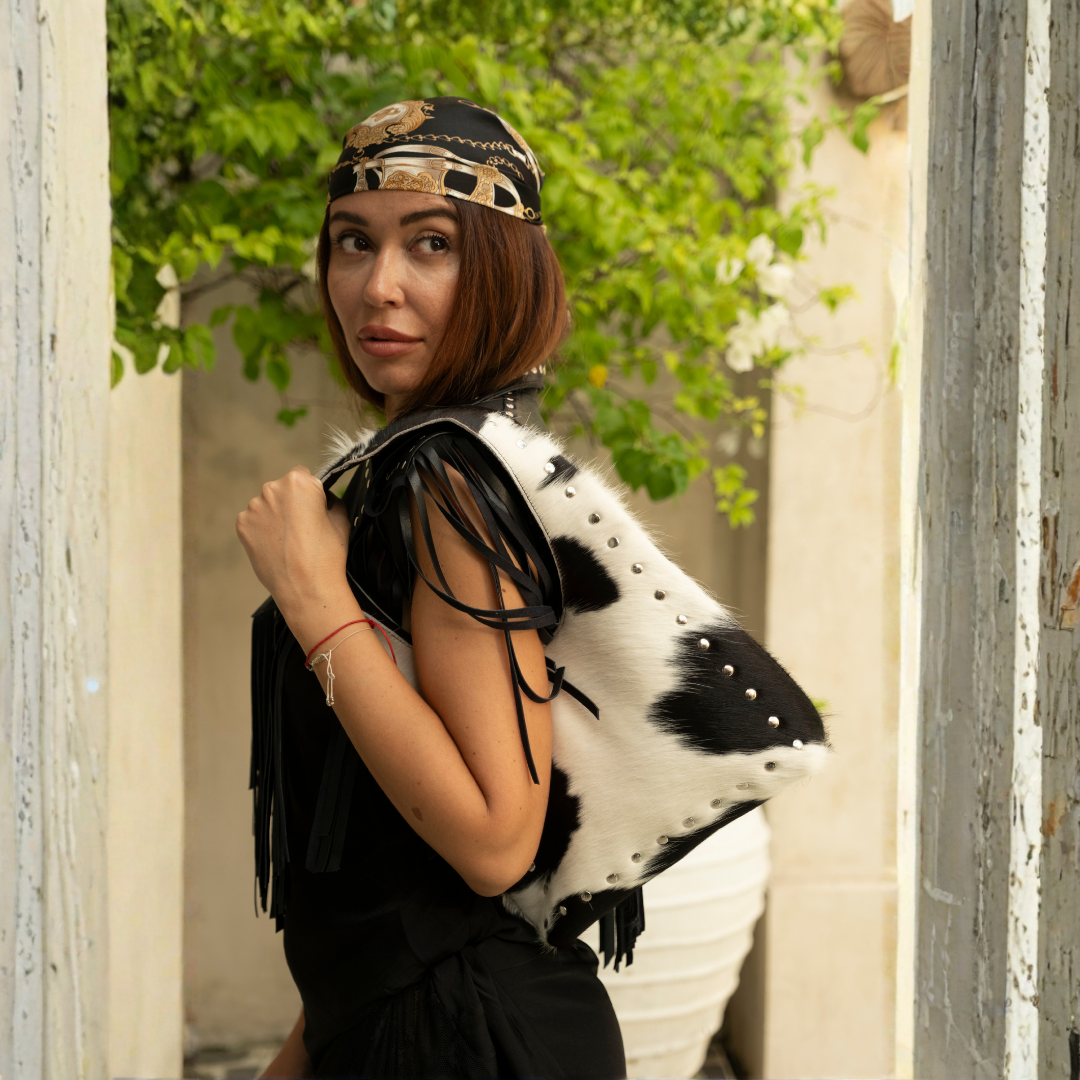 Wrangler Cowhide Bag from House of Hide in black and white cow-print cowhide leather being worn as shoulder bag by model against white wall with green climbing ivy