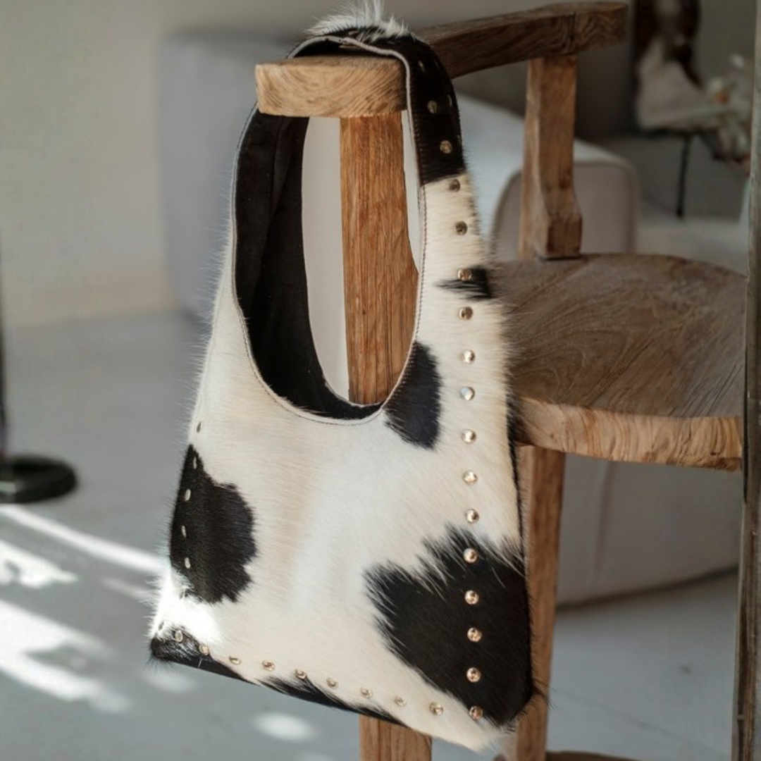 Wrangler Cowhide Bag from House of Hide in black and white patchwork cowhide leather with gold-tone studs being displayed hanging on rustic wooden chair in softly lit indoor setting